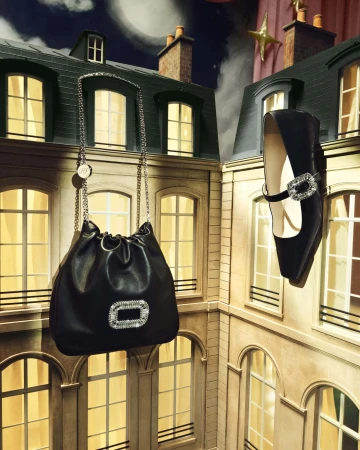 Elevated view of a black bucket bag with a chain strap and a black, jeweled shoe, suspended in front of illuminated building windows.