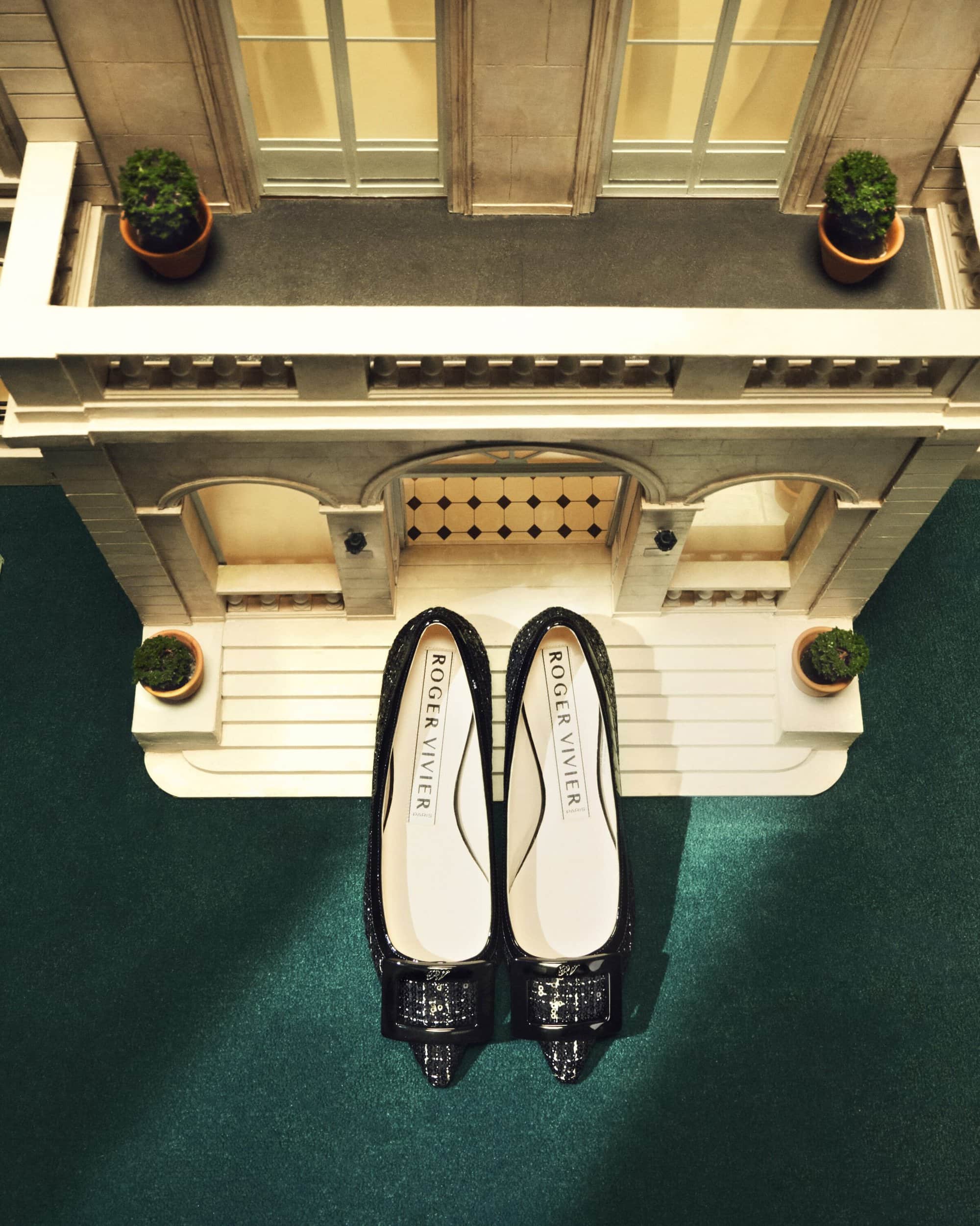 Elevated view of Roger Vivier flats in front of a miniature building facade with potted plants on balconies on a green surface.