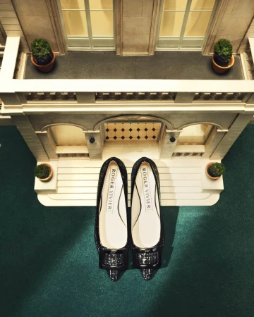 Elevated view of Roger Vivier flats in front of a miniature building facade with potted plants on balconies on a green surface.