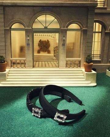 Two black embellished headbands rest on green fabric, with a miniature building with steps in the background. Fashion accessory shot.