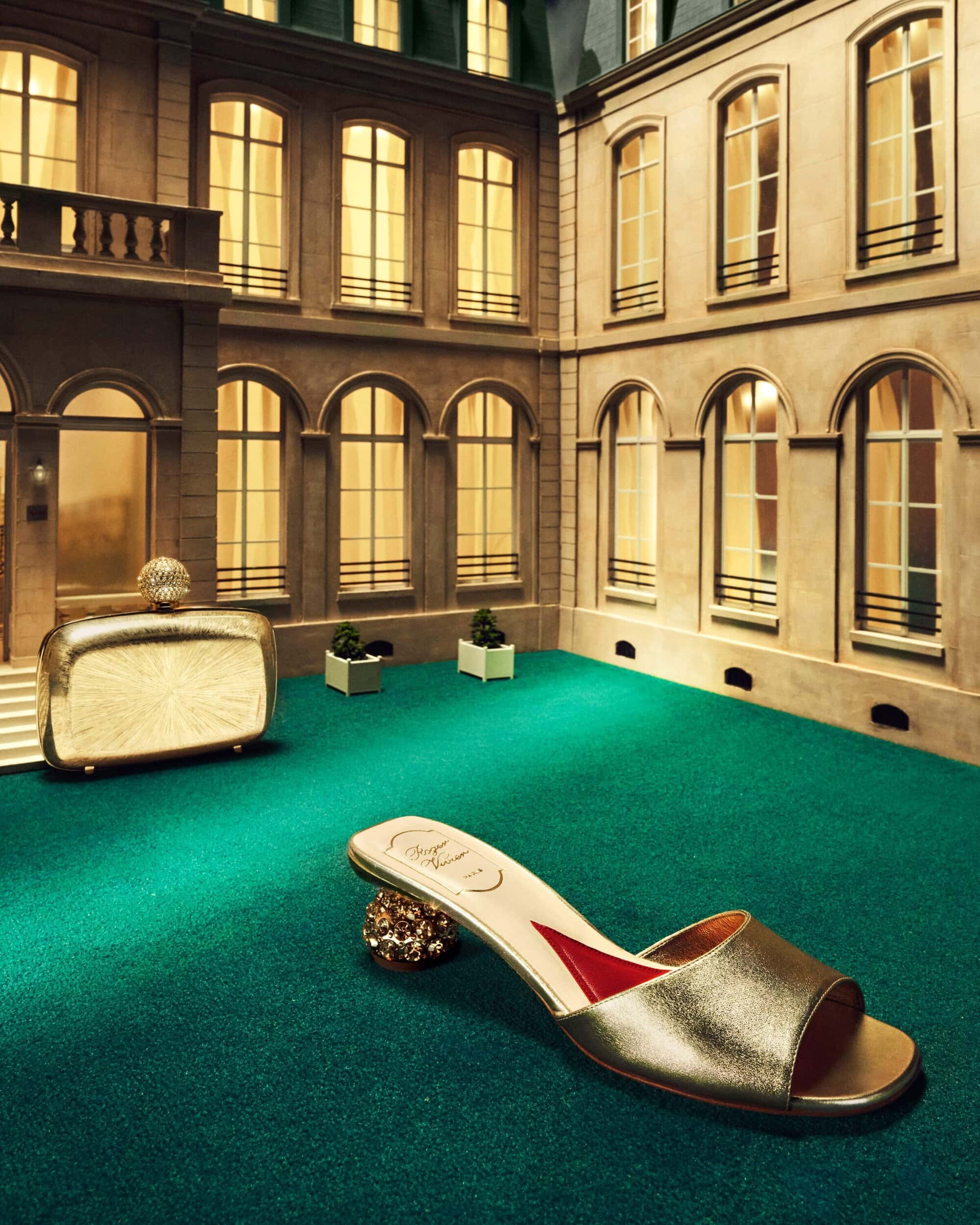 Gold high-heeled mule sits on a green surface with a matching clutch in the background against a backdrop of a light-colored building.