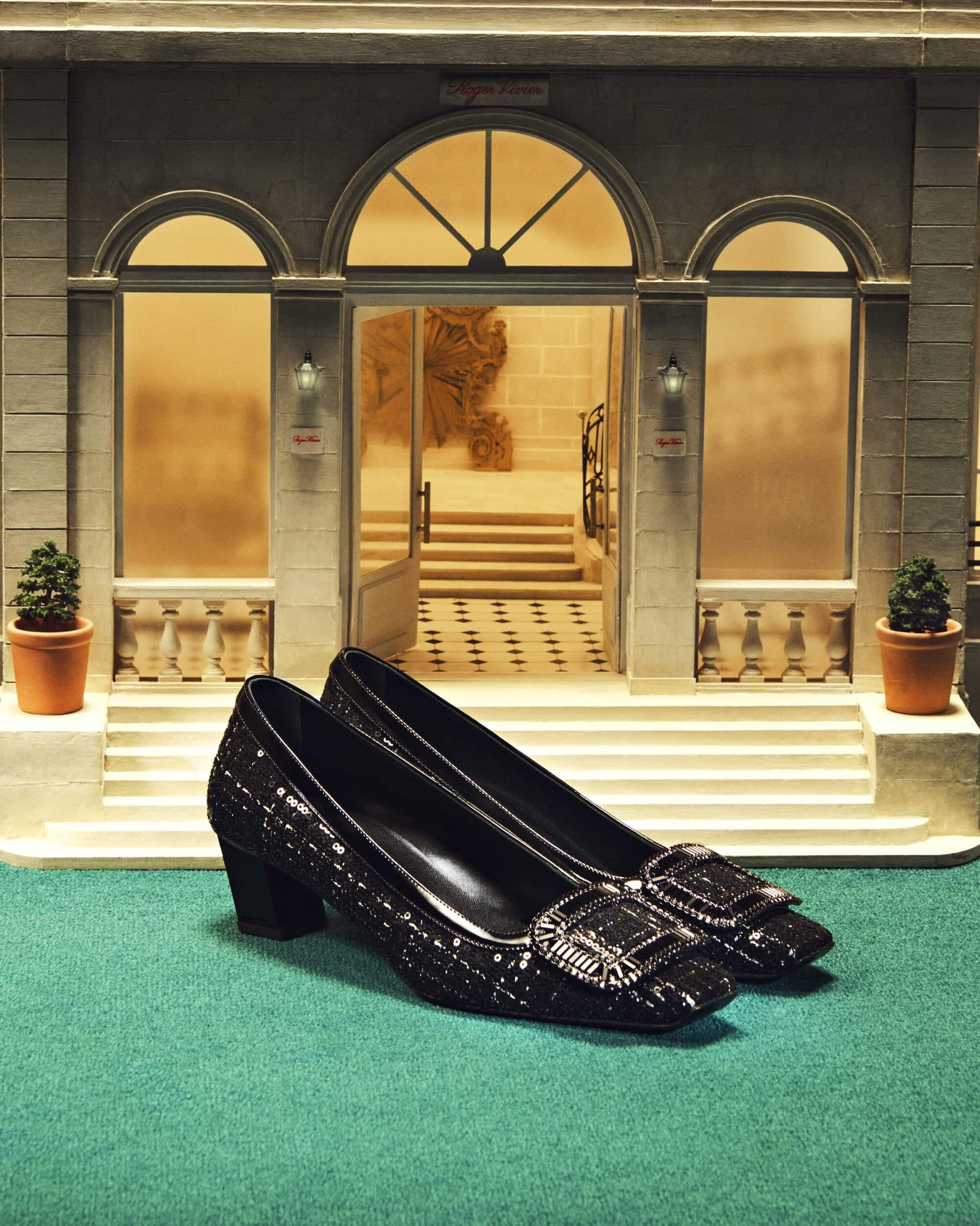 Elegant black heeled shoes with decorative buckles on a green surface, presented in front of a detailed miniature building facade.