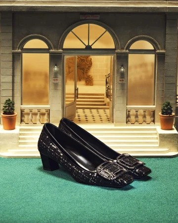 Elegant black heeled shoes with decorative buckles on a green surface, presented in front of a detailed miniature building facade.