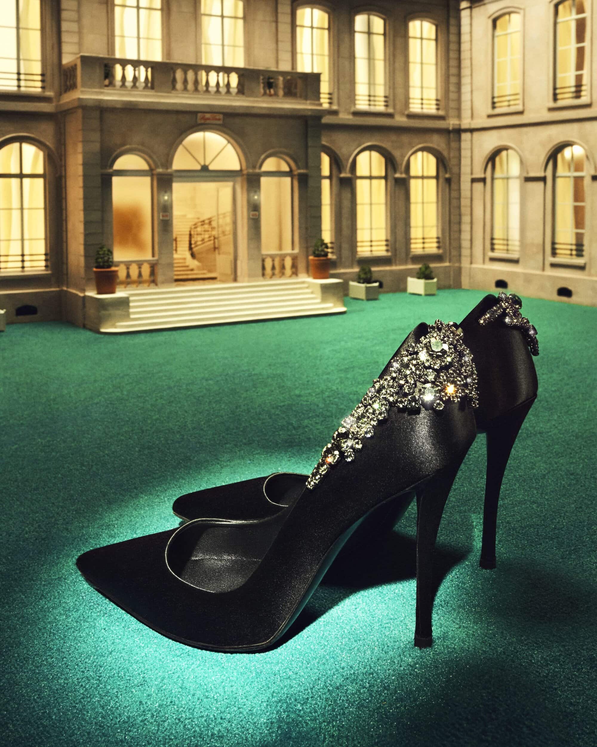 Elegant black satin high-heeled shoes embellished with crystals displayed on a green surface against a building backdrop.