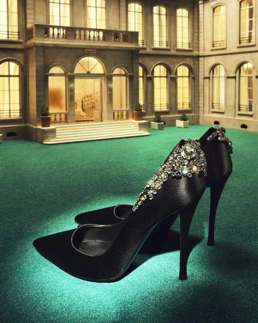Elegant black satin high-heeled shoes embellished with crystals displayed on a green surface against a building backdrop.