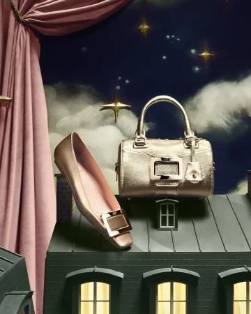 Elevated view of a woman's gold ballet flat and a gold handbag, staged on a miniature building set against a backdrop of pink curtains and a night sky.