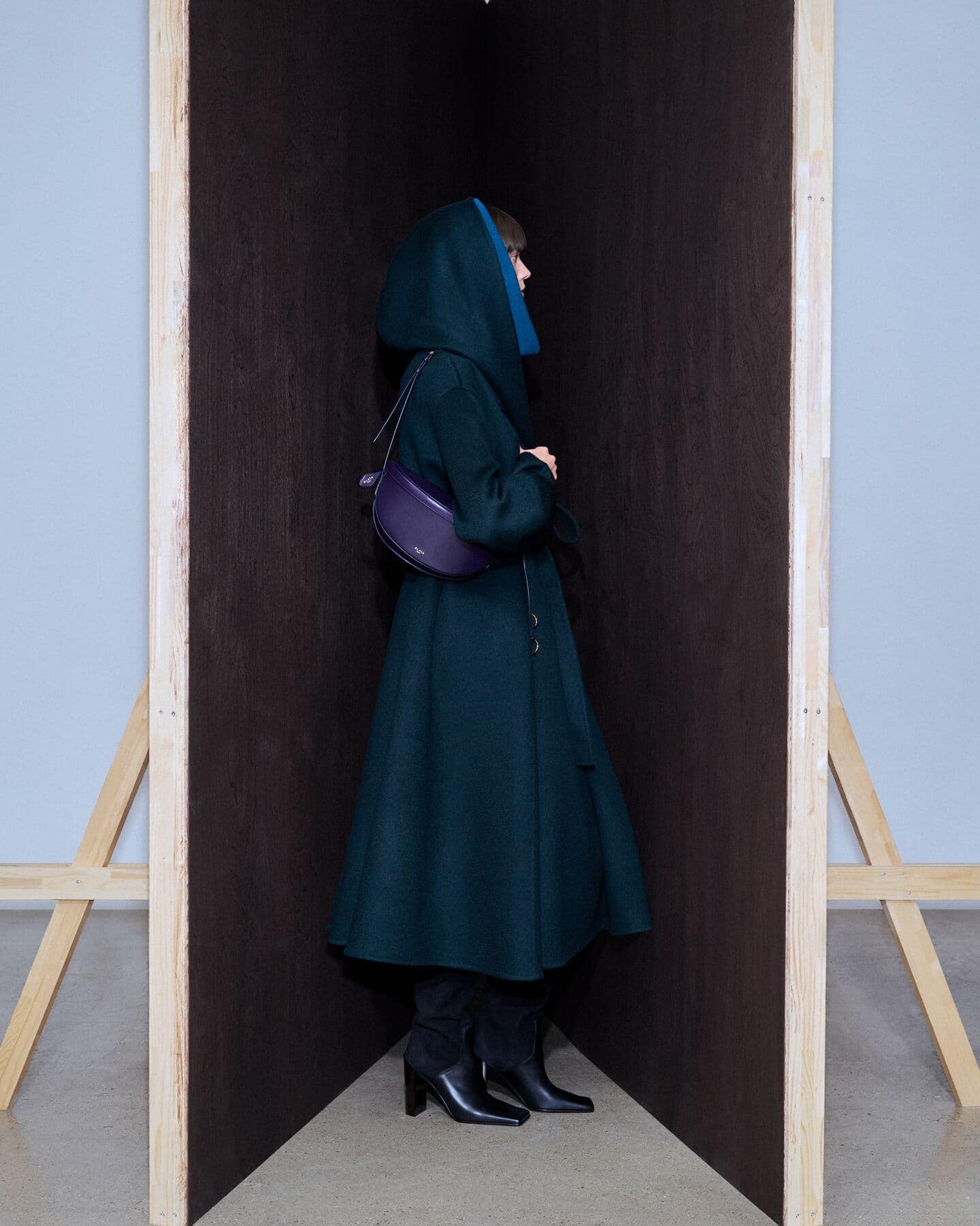 A stylish adult female stands in an enclosed space wearing a green hooded coat, black boots, and a purple purse in an indoor setting.