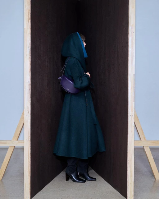 A stylish adult female stands in an enclosed space wearing a green hooded coat, black boots, and a purple purse in an indoor setting.