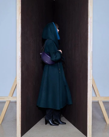 A stylish adult female stands in an enclosed space wearing a green hooded coat, black boots, and a purple purse in an indoor setting.