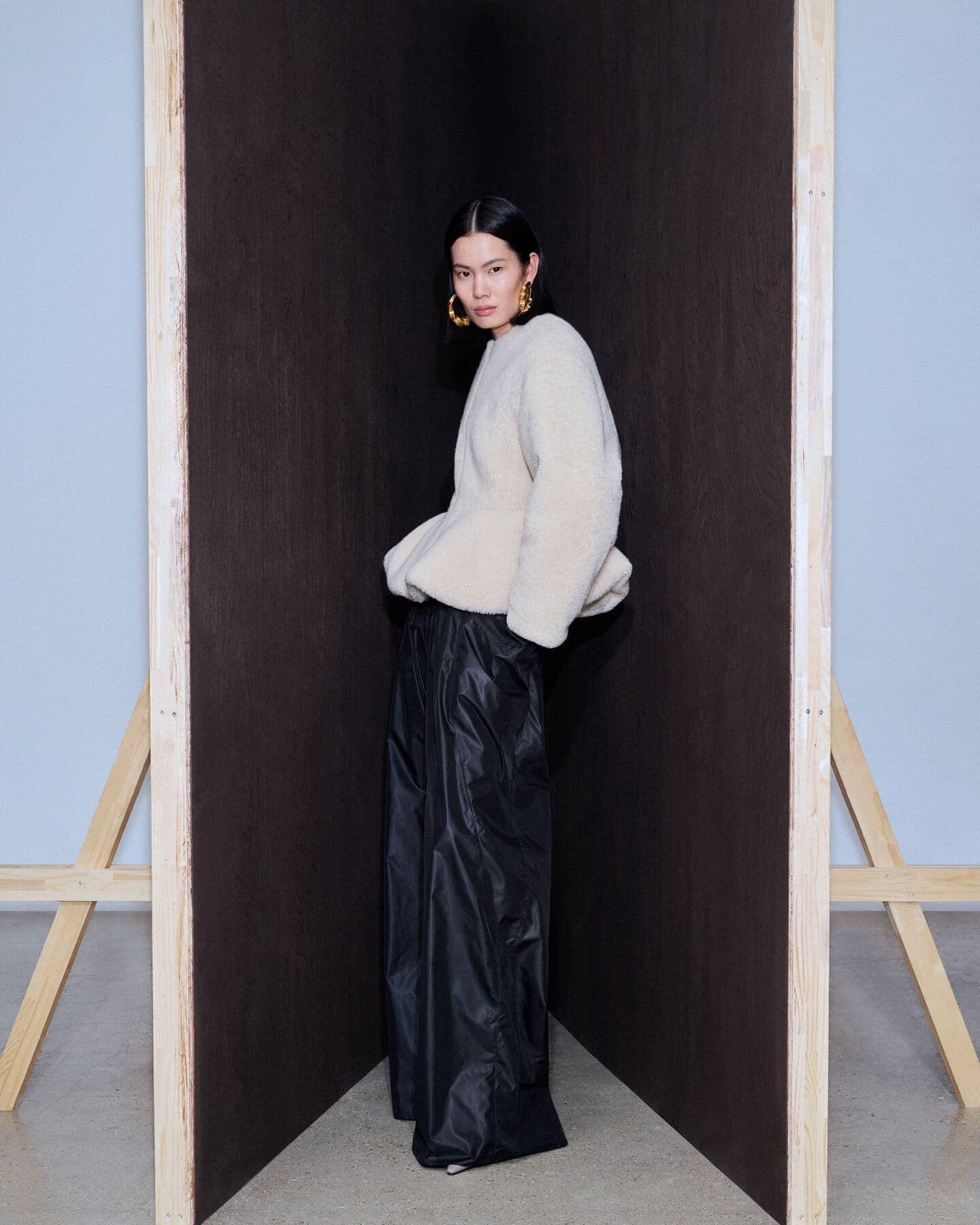 Asian woman poses in a fashion shot wearing a beige top and black pants in an indoor studio with wood and light-blue backdrop.