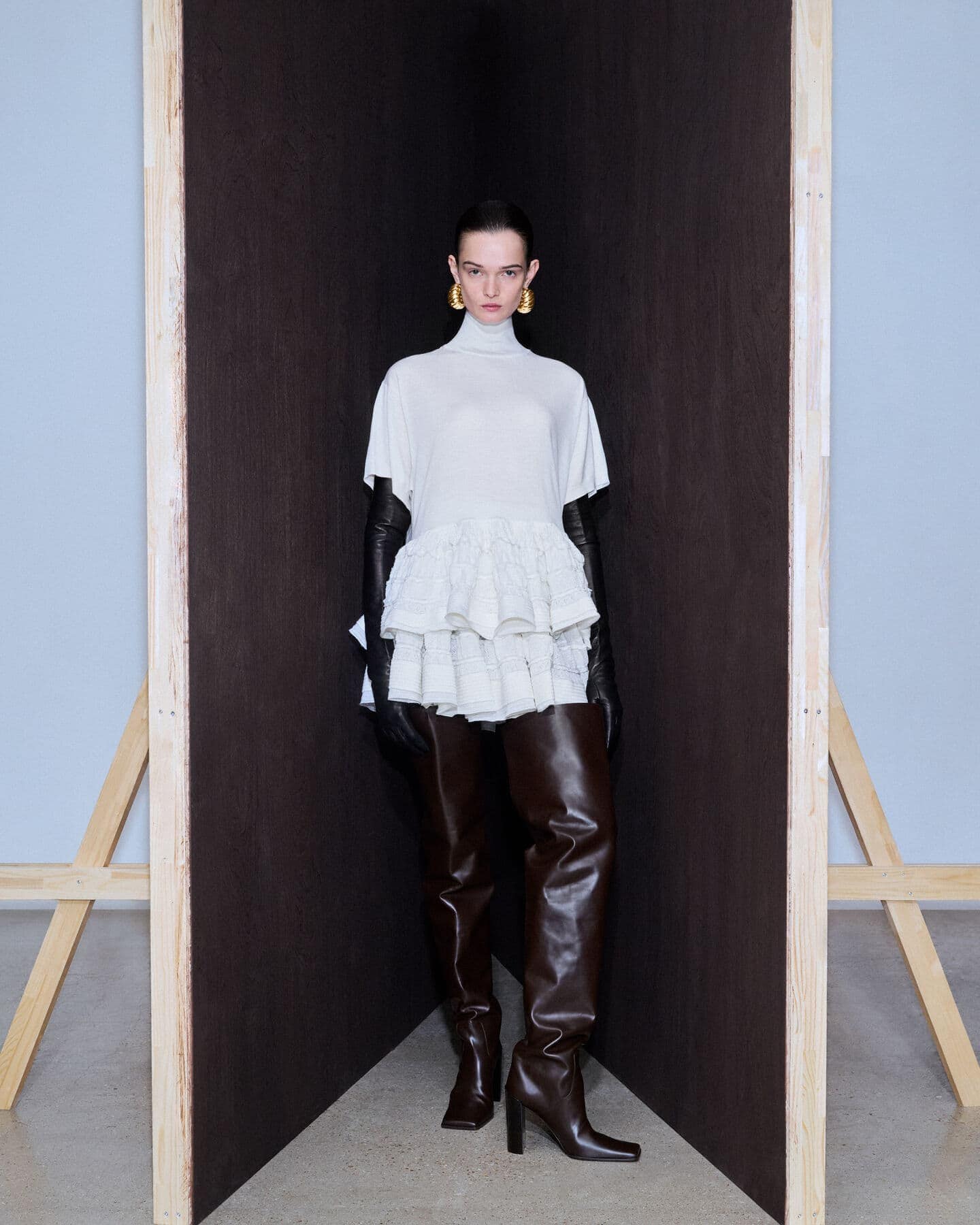 A woman in a white top, black gloves, and brown thigh-high boots stands in a studio setting framed by a wooden structure.