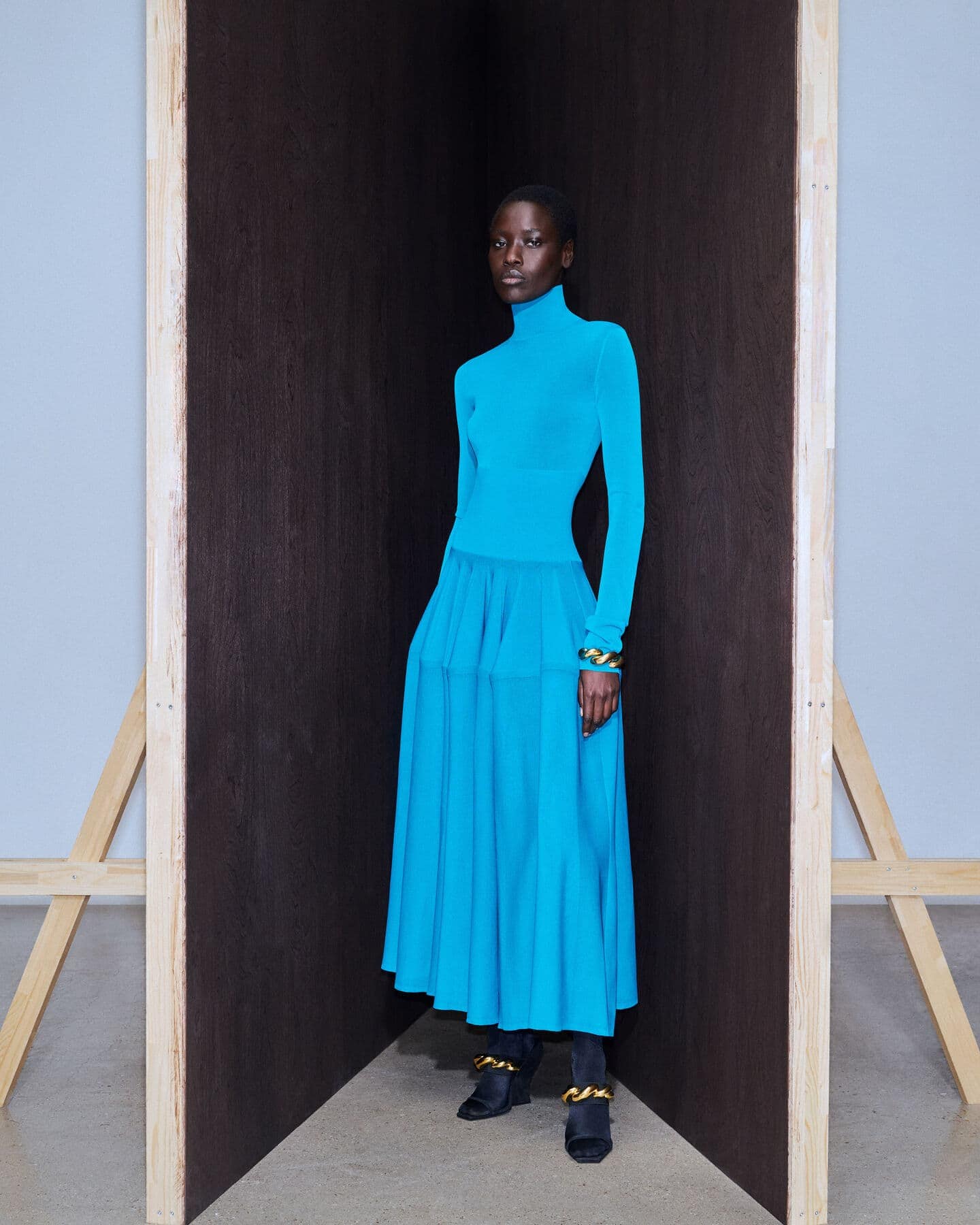 An elegant Black woman poses in a bright-blue turtleneck dress with long sleeves, gold bangles, and black shoes in a minimal photo studio.