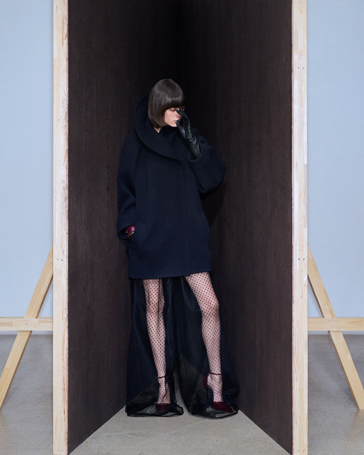 A woman posing in a dark coat and polka dot tights stands in a wooden structure in a studio. Fashion editorial style.