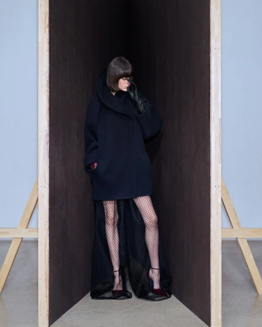 A woman posing in a dark coat and polka dot tights stands in a wooden structure in a studio. Fashion editorial style.
