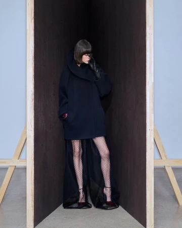 A woman posing in a dark coat and polka dot tights stands in a wooden structure in a studio. Fashion editorial style.