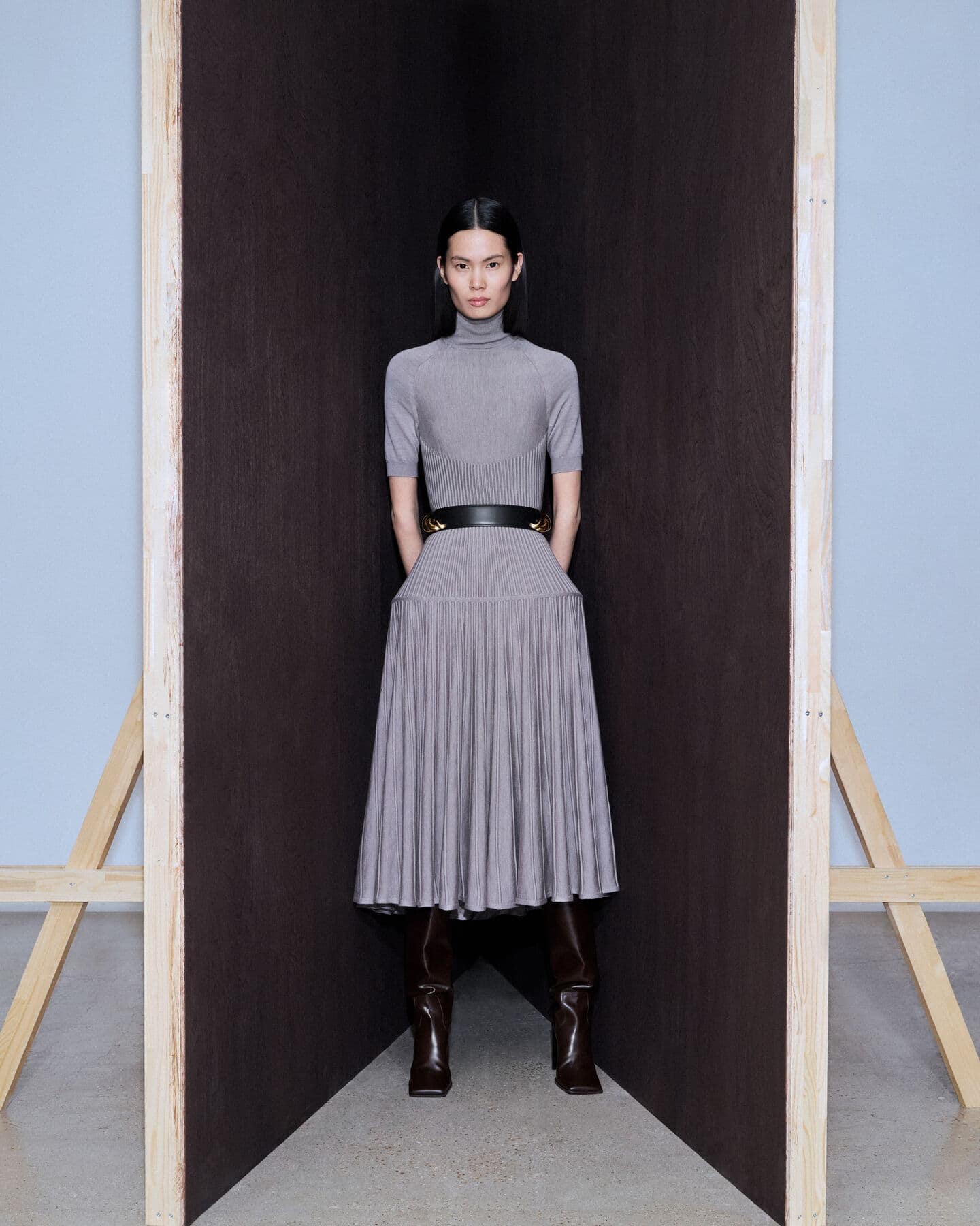 Full-length shot of a woman in a grey ribbed, pleated dress and brown boots, standing in front of a dark panel with a light blue background.