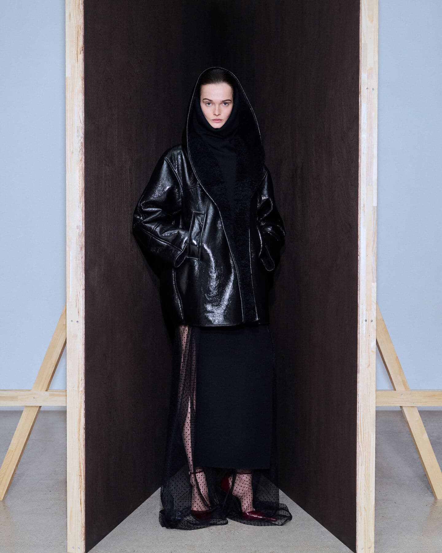 A woman in a hooded black coat and black dress stands in a framed indoor setting, showcasing a modern fashion aesthetic and neutral tones.