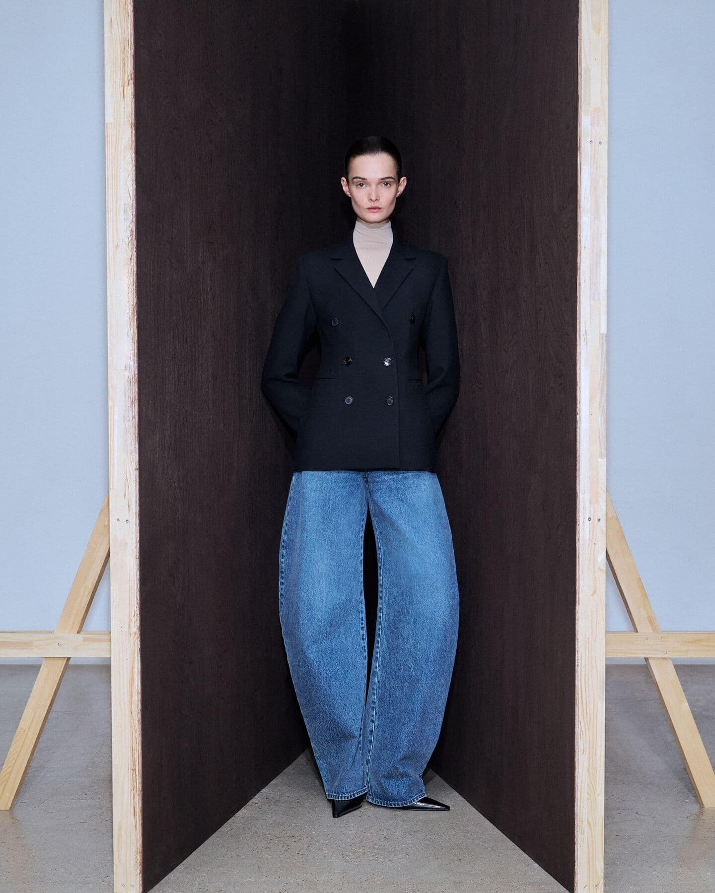 Fashion model in oversized denim and a black blazer poses against a dark backdrop with natural light and a minimalist aesthetic.