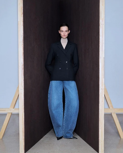 Fashion model in oversized denim and a black blazer poses against a dark backdrop with natural light and a minimalist aesthetic.