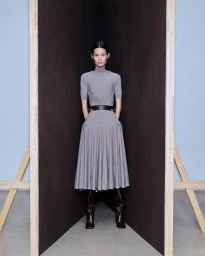 Model wearing a grey turtleneck midi dress and dark boots standing between angled dark panels