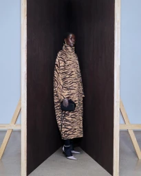 Model standing inside a dark wooden corner structure wearing a tiger-print coat and holding a black handbag