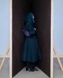 Model wearing a dark green hooded coat and carrying a purple handbag standing in a wooden corner structure for Alaïa