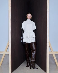 Model standing inside a dark corner structure wearing a white ruffled top and tall brown leather boots