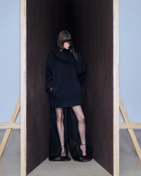 Model in dark hooded coat and polka dot tights posing within a wooden architectural corner structure