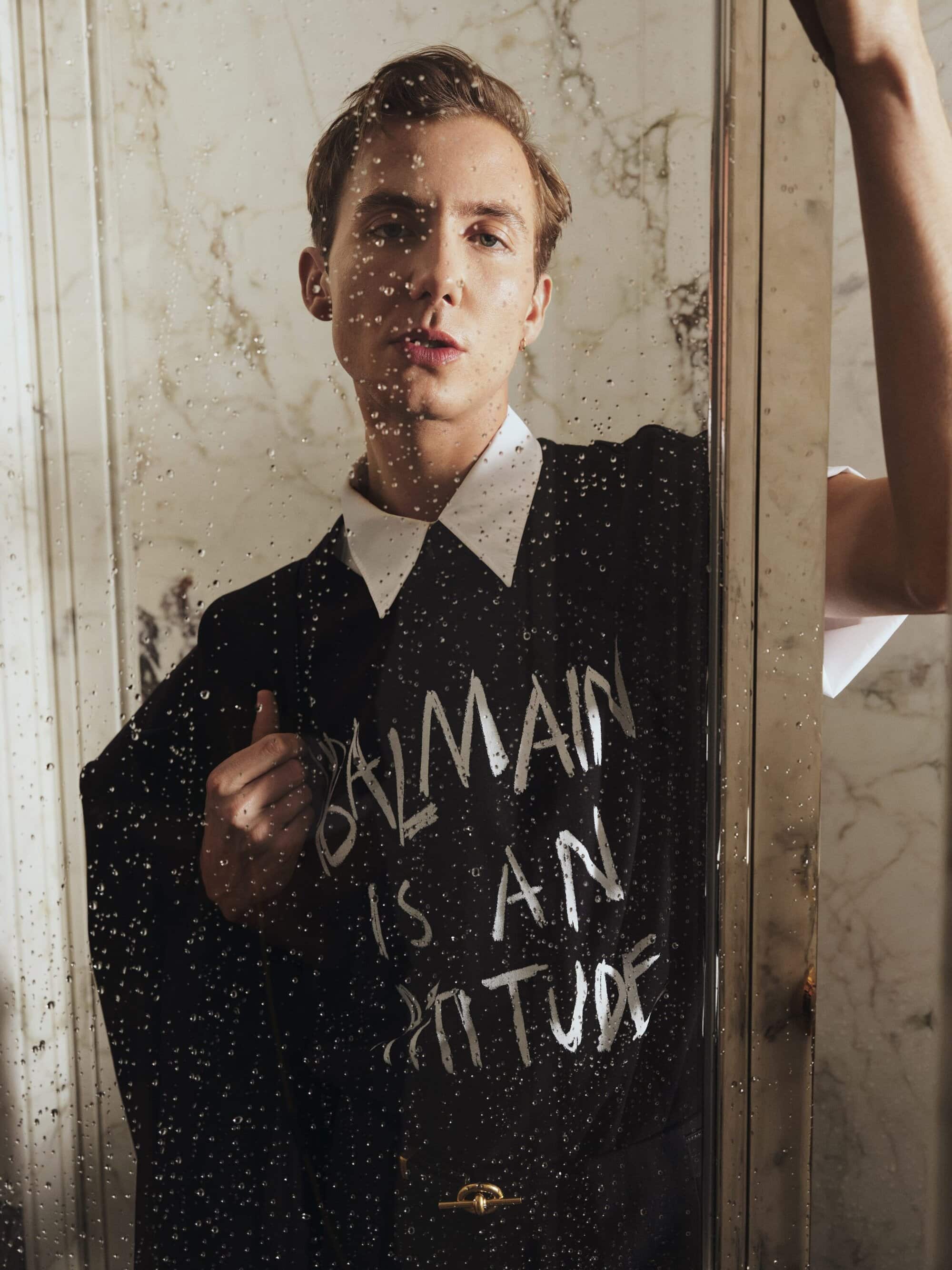 Fashion portrait of a male model wearing a black shirt with white text, posing in front of a marble wall with a reflective glass door.