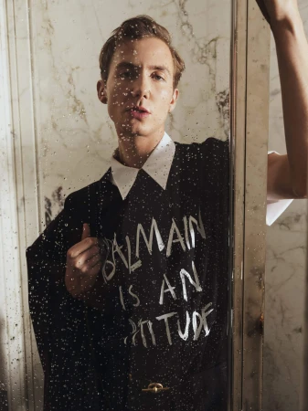 Fashion portrait of a male model wearing a black shirt with white text, posing in front of a marble wall with a reflective glass door.
