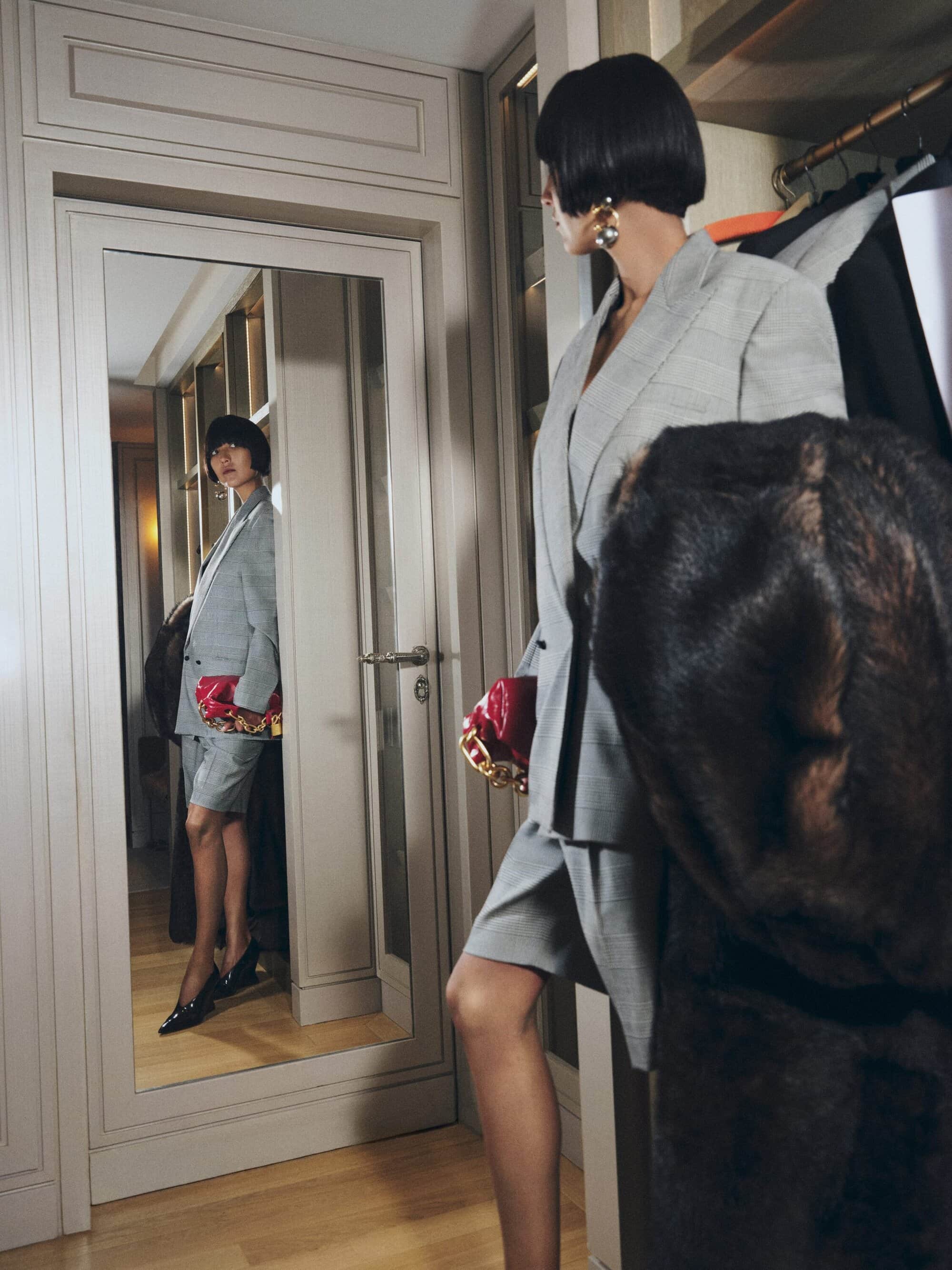 An adult female in a grey suit stands in front of a mirror indoors, with a pink purse and a fur coat nearby, fashion editorial.