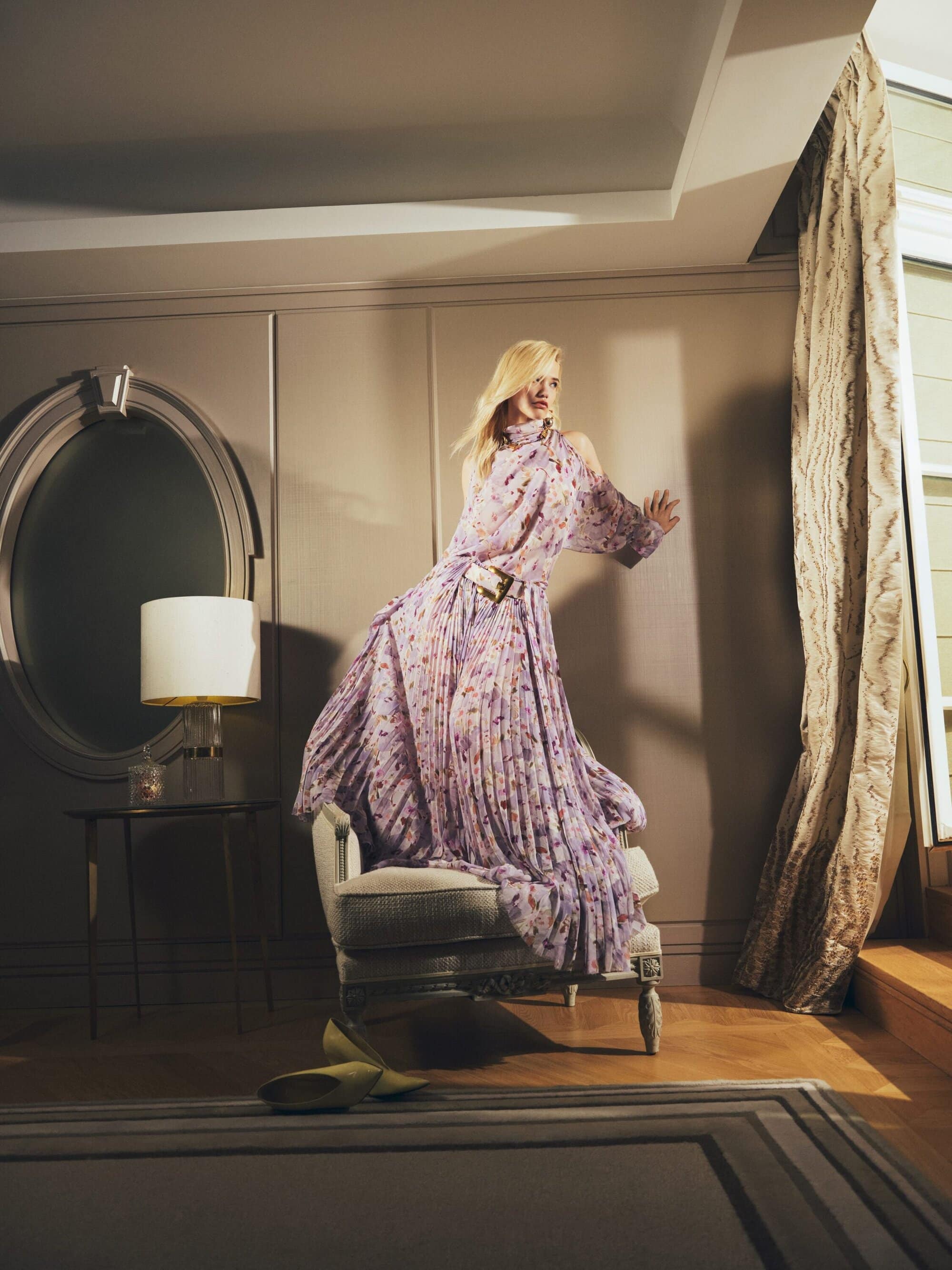 Woman in floral dress posing in an interior. Dress flows as she raises one arm to touch a wall, with chair and shoes.