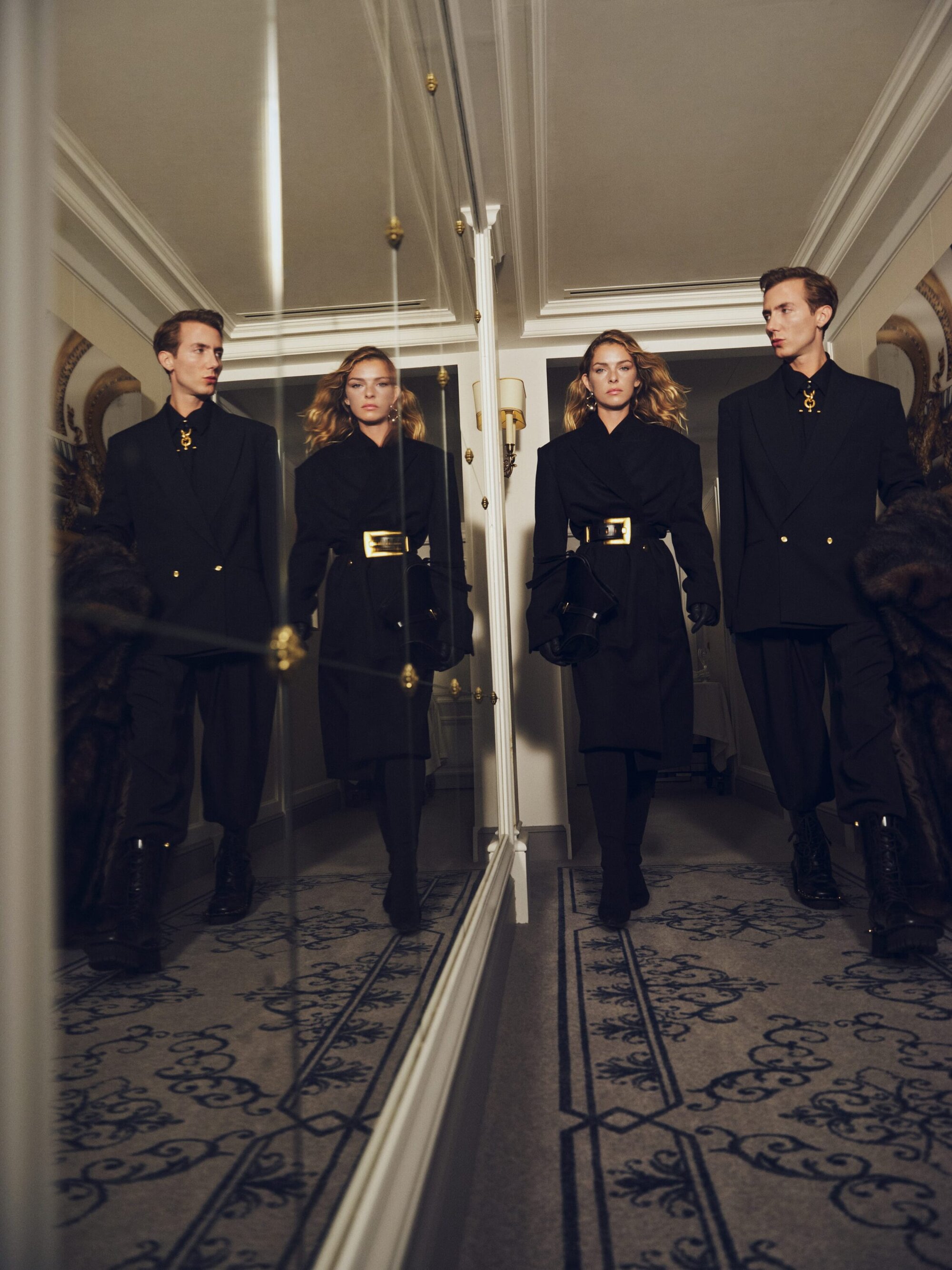 Two fashion models in elegant black coats walk in a brightly lit hallway with a mirror reflecting their images, creating a symmetrical scene.