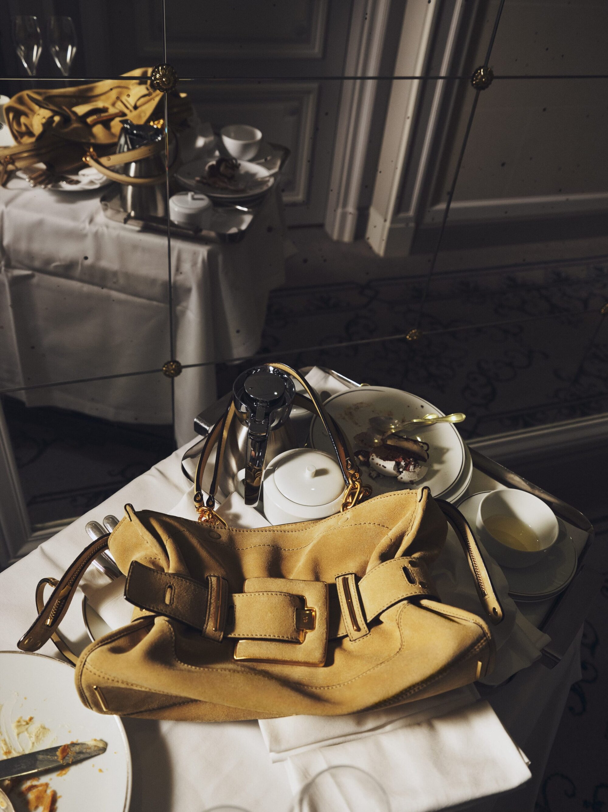Beige suede handbag displayed on a white tablecloth next to tableware, reflected in a mirror creating a luxurious, stylish, and high-end fashion vibe.