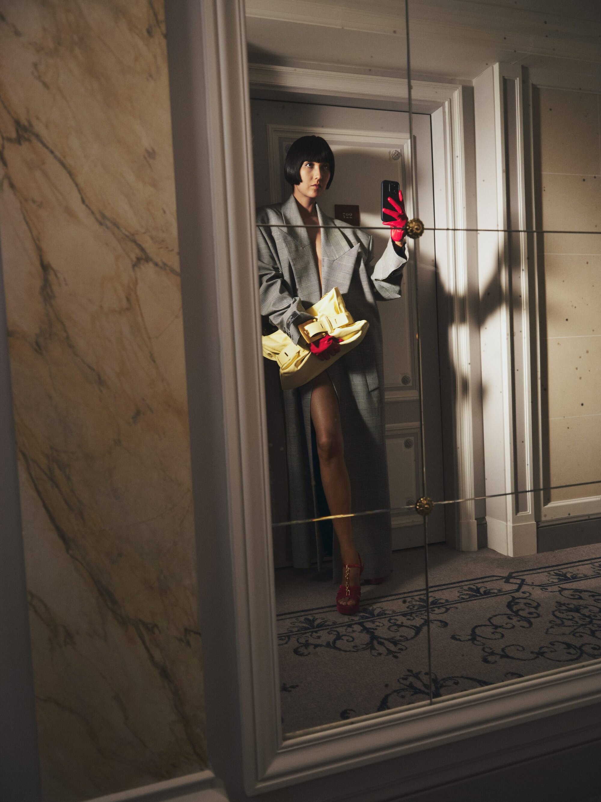 A woman with bobbed hair poses for a selfie in a mirror, wearing a grey coat, red gloves, and carrying a yellow bag.