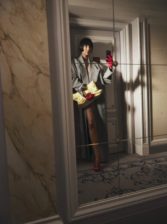 A woman with bobbed hair poses for a selfie in a mirror, wearing a grey coat, red gloves, and carrying a yellow bag.