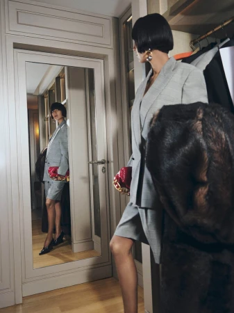 Model in a grey suit holding a red handbag standing in a mirrored walk-in closet for Balmain Holiday 2025 campaign.