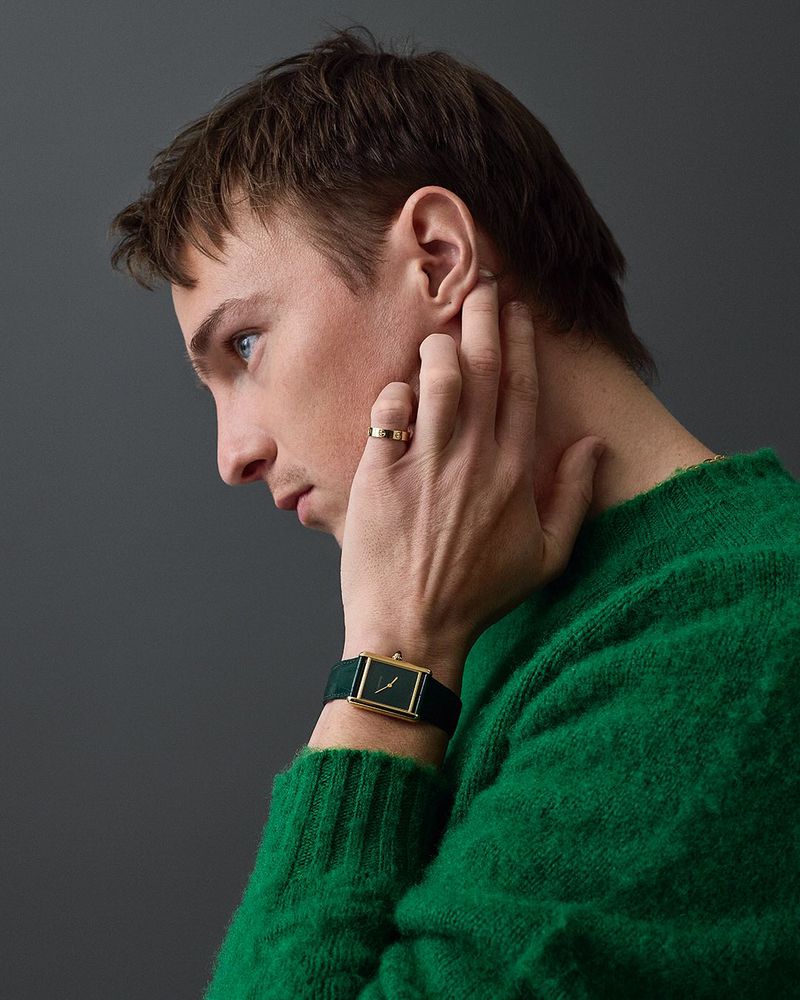 Man in green sweater wearing gold ring and wristwatch against grey background in studio portrait emphasizing fashion and style.