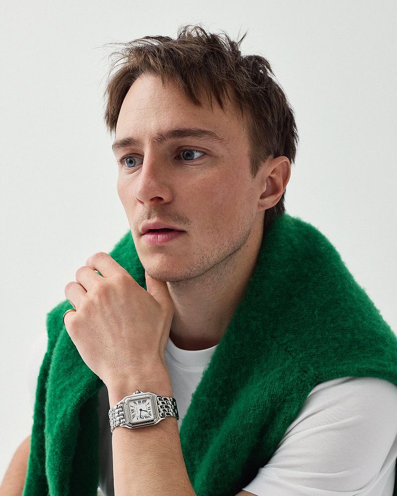 Close-up of a dark-haired man wearing a green sweater and a silver watch, posing in front of a clean, white background.