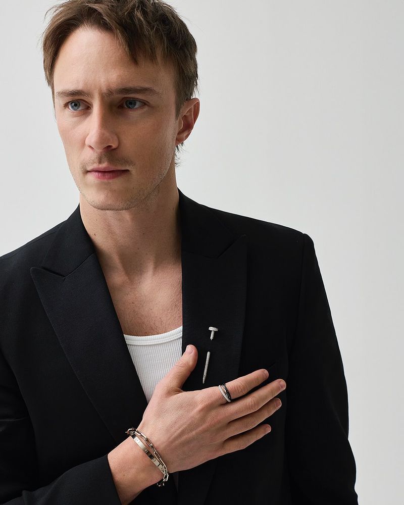 Close-up studio shot of a fair-skinned male adult in a black suit jacket and white undershirt, adorned with jewelry and a nail pin.