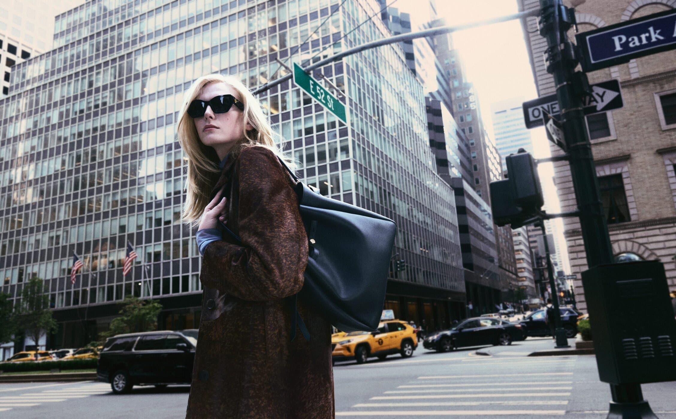 Woman in coat and sunglasses carrying a tote bag on a city street with skyscrapers and traffic signals, capturing a lifestyle moment.
