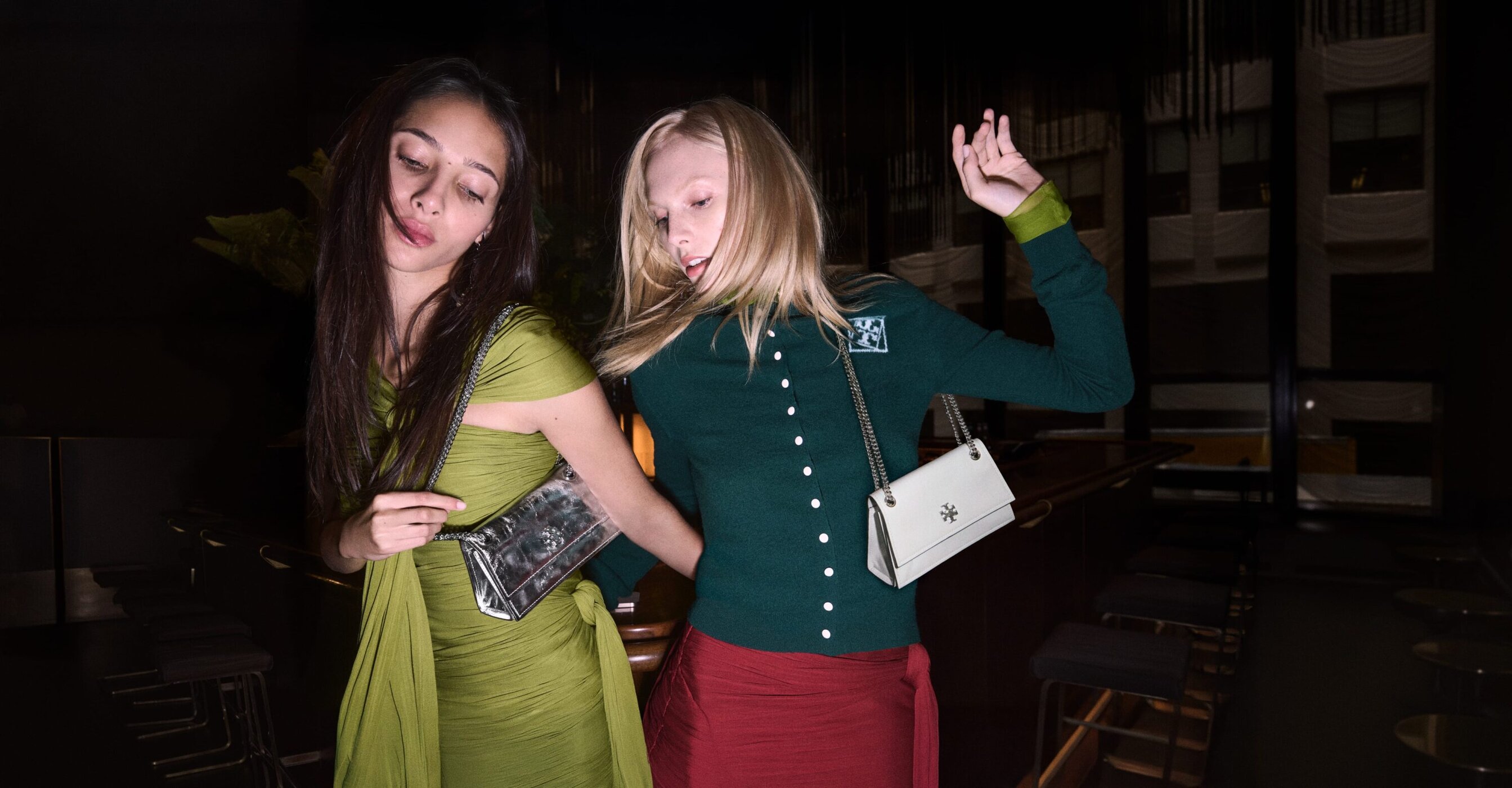 Two fashion models pose indoors with handbags. One woman wears a green dress; the other is in a green cardigan and red skirt.