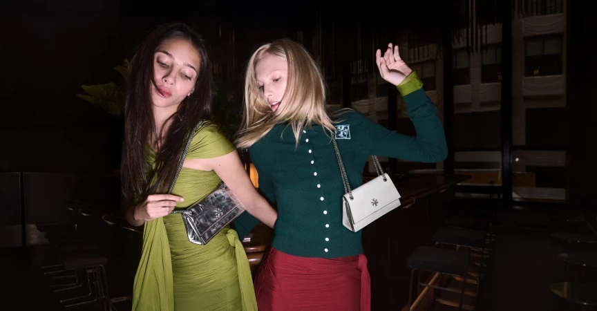 Two fashion models pose indoors with handbags. One woman wears a green dress; the other is in a green cardigan and red skirt.