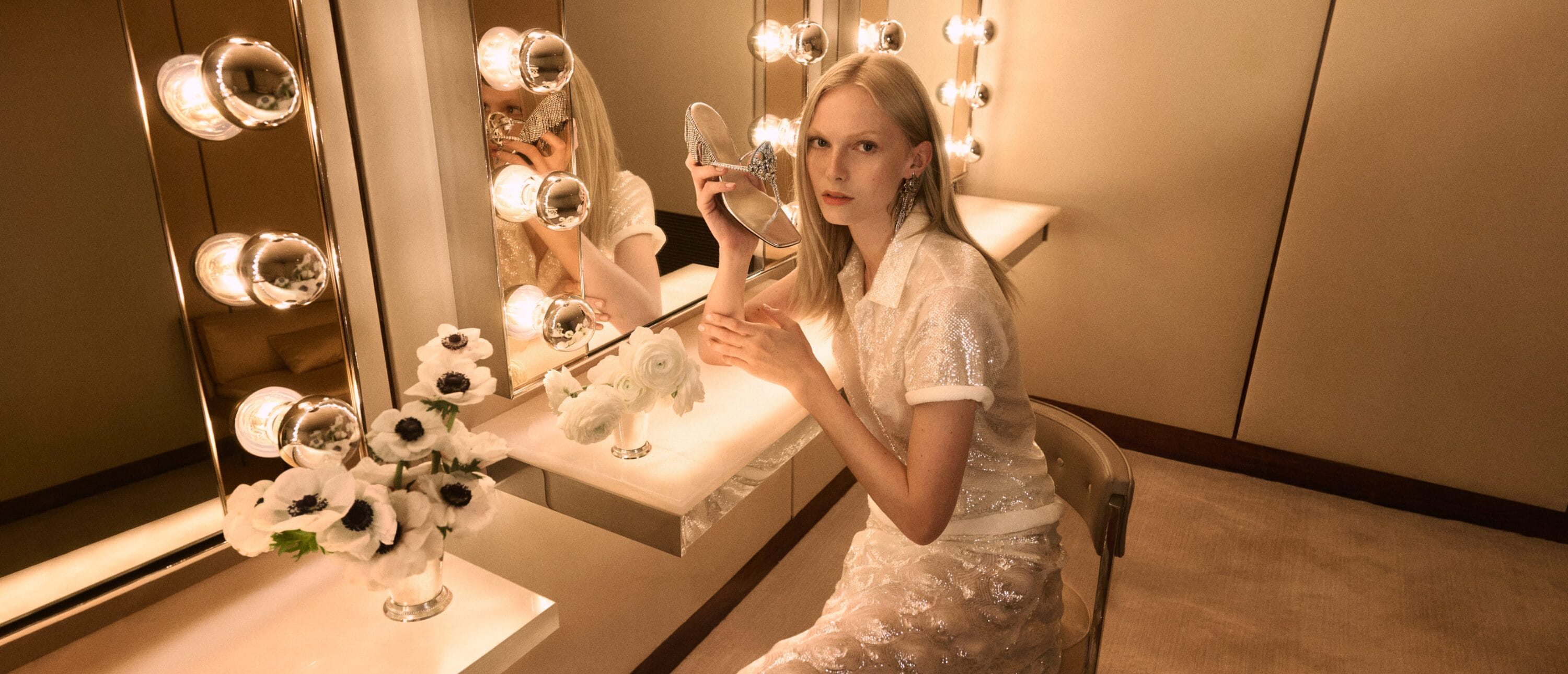 Woman poses seated at lighted vanity wearing a sequined top and skirt, holding a silver shoe indoors with white flower bouquets.