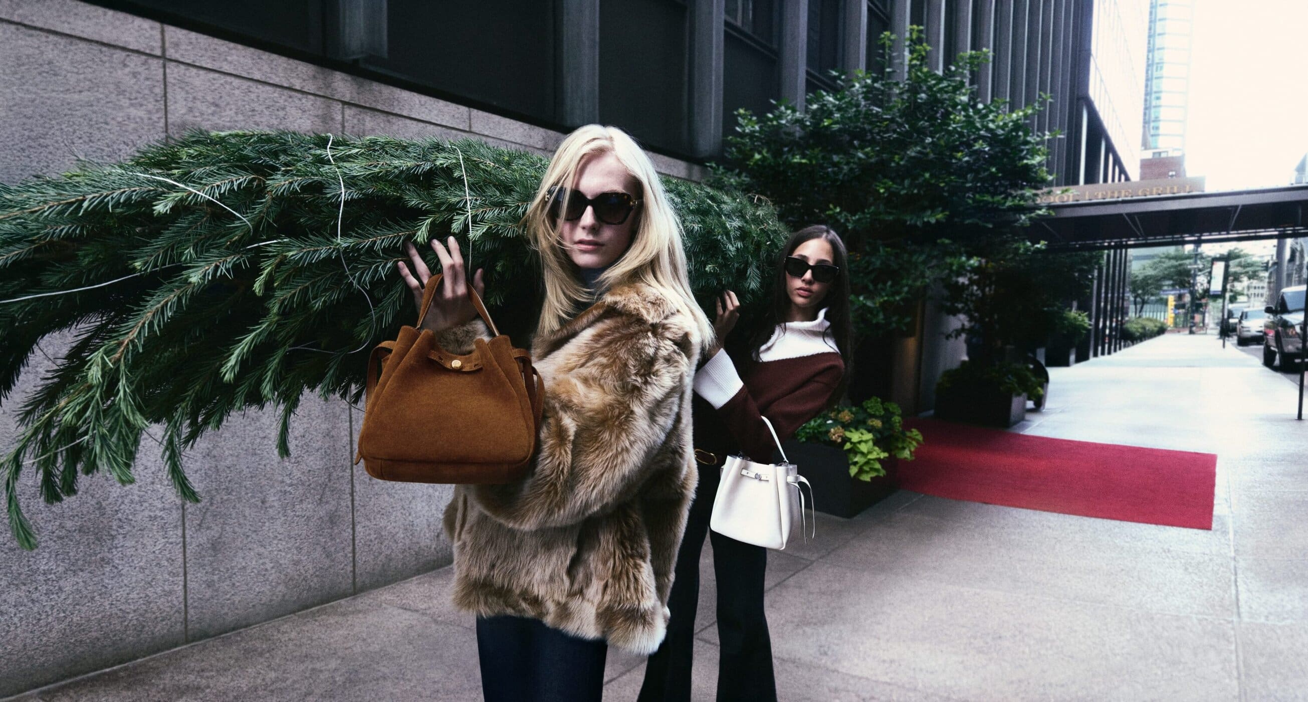Two women walk on an urban street in sunglasses carrying a small brown purse, and one also has a small Christmas tree.