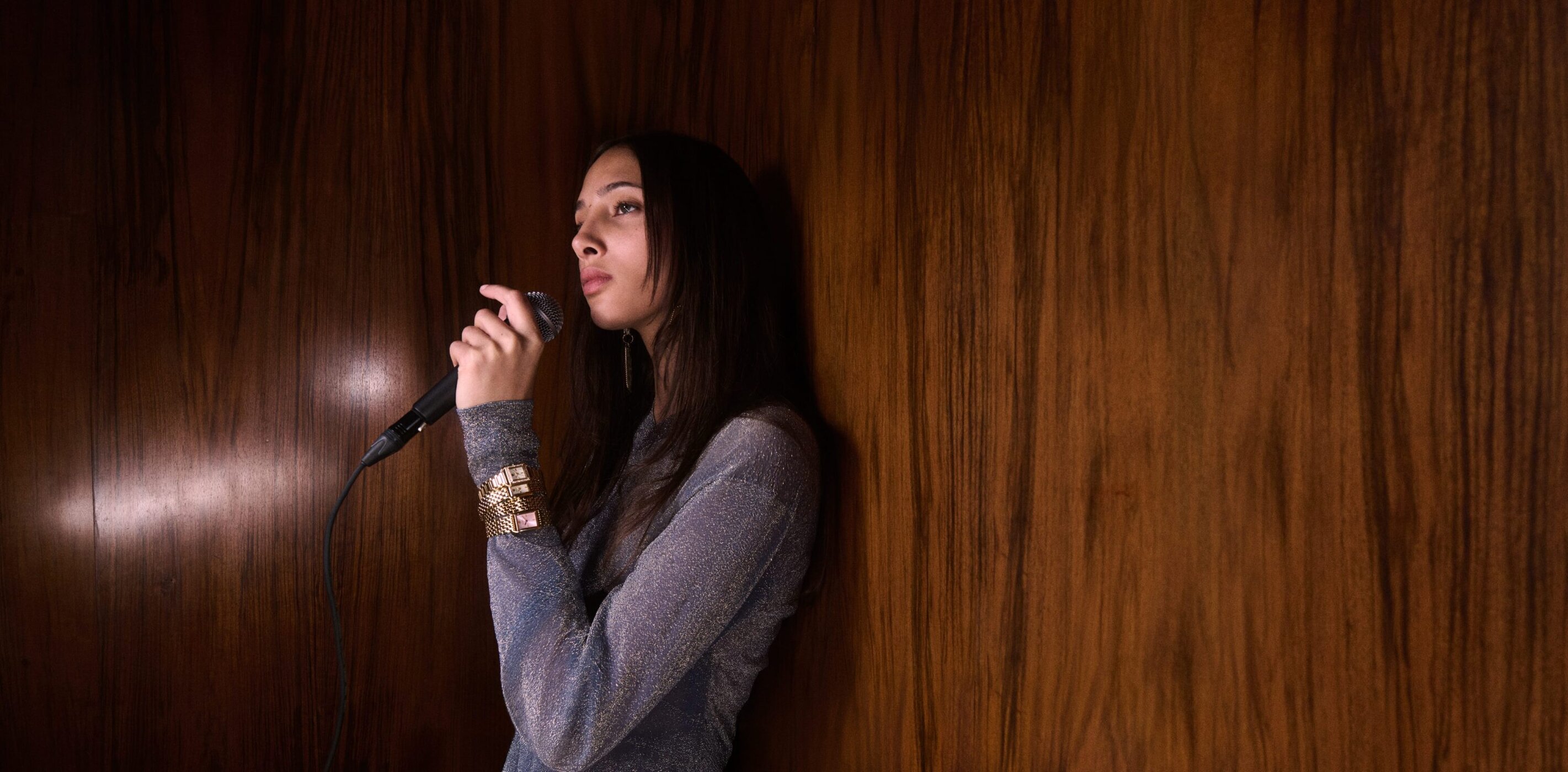 Woman sings on microphone indoors with wood background. Woman wearing sparkling dress and metallic-gold cuff bracelet.
