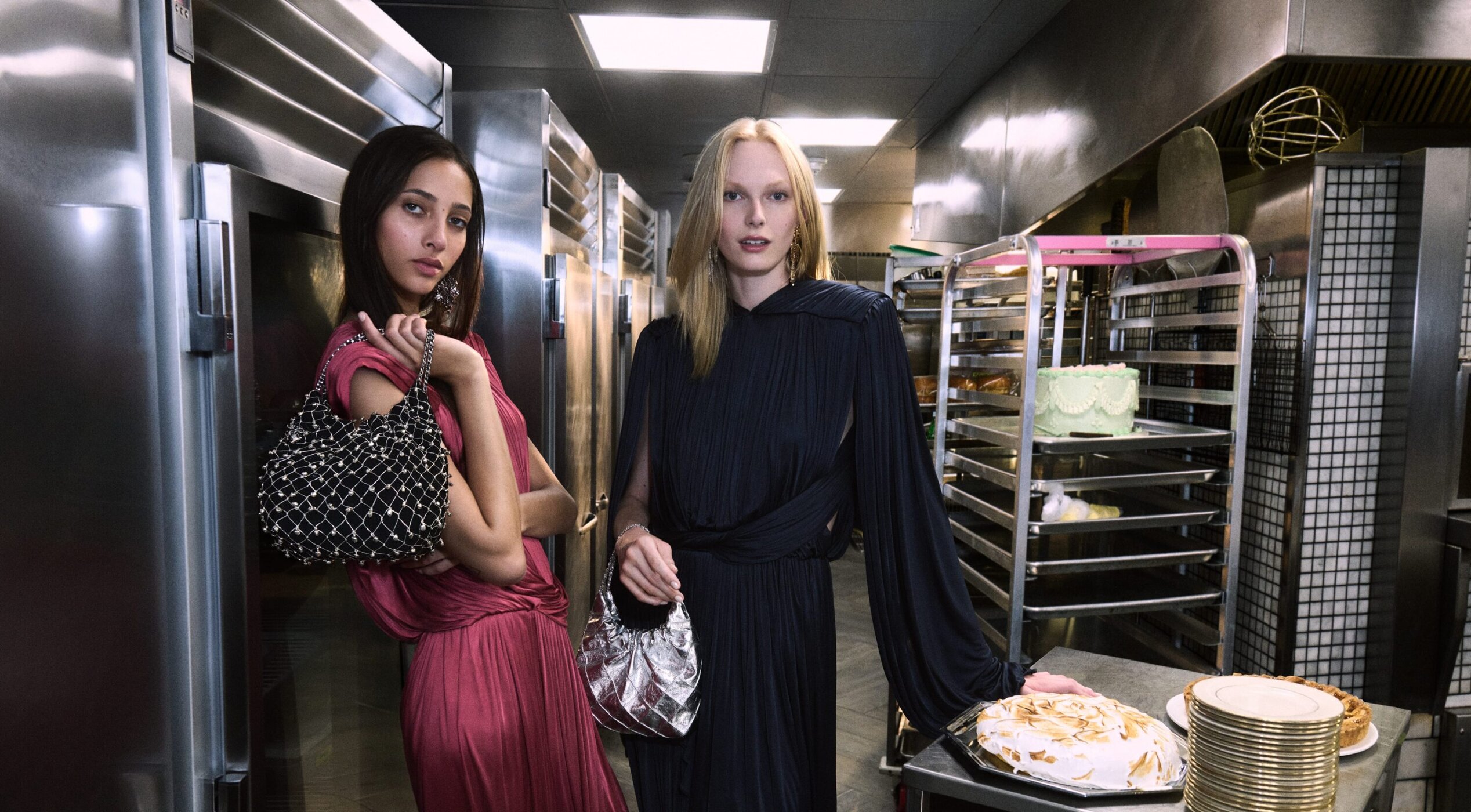 Two women in formal dresses hold purses in a commercial kitchen with stainless steel appliances and desserts on racks and counters.