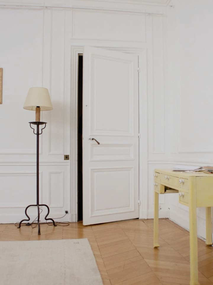 Interior view of a room featuring an open white door, lamp with a beige shade, and a small desk, all set against white walls.