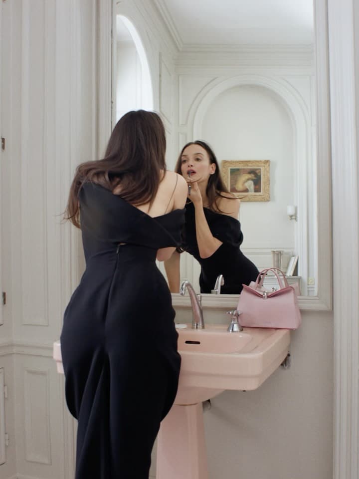 A woman in a black dress applies lipstick while looking in a mirror in a bathroom with pink sink, with a small pink handbag.