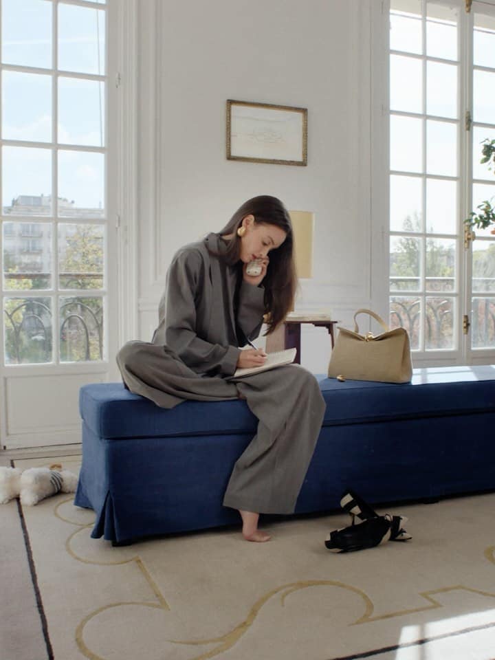 A woman in a grey suit sits cross-legged on a blue bench in a bright room, writing in a notebook while on the phone.